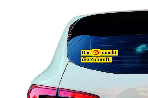 Ein Aufkleber auf der Heckscheibe eines weißen Autos zeigt den Schriftzug „Das E macht die Zukunft.“ Das „E“ ist als rotes Symbol in einem gelben Kreis gestaltet. Die Schrift ist in schwarzer Farbe auf gelbem Hintergrund gedruckt.